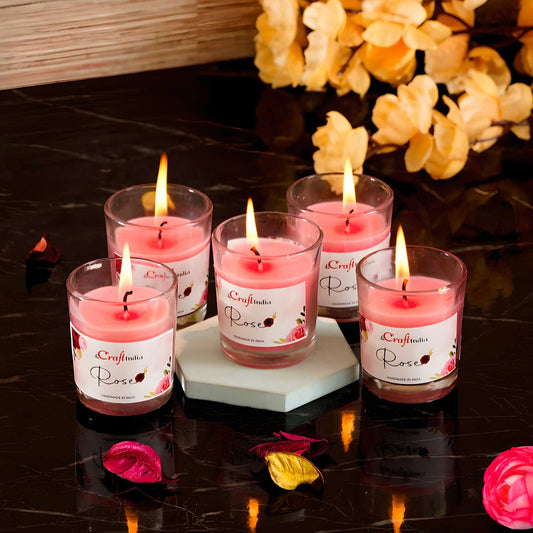Set of six pink candles in glass holders with visible flames on a dark surface, surrounded by decorative flowers.